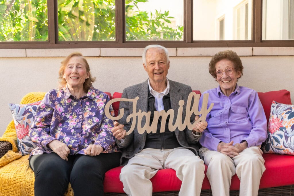 Three elderly people sitting on a couch holding a sign that says family.